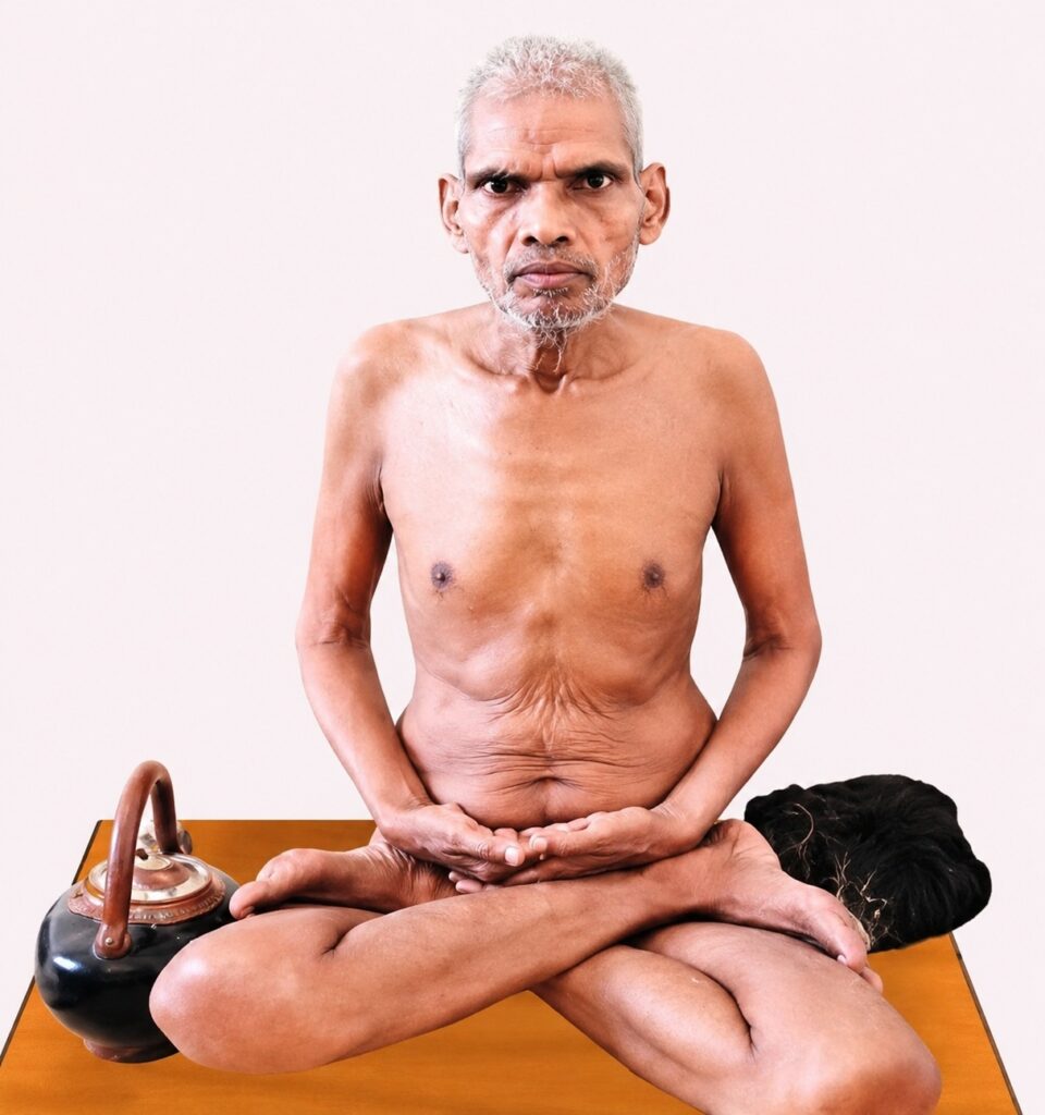 Older shirtless man with gray hair sits cross-legged on a wooden table, meditating with hands resting in lap; a kettle sits beside him on the table.