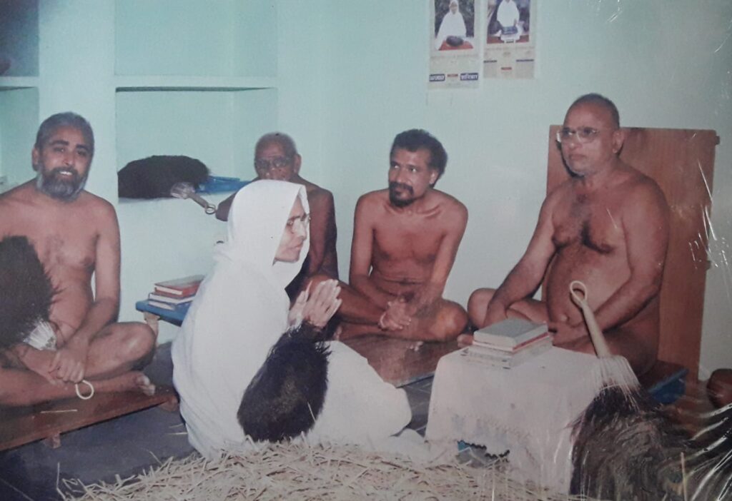 A nun in a white habit praying with folded hands, seated among shirtless men in a simple room.