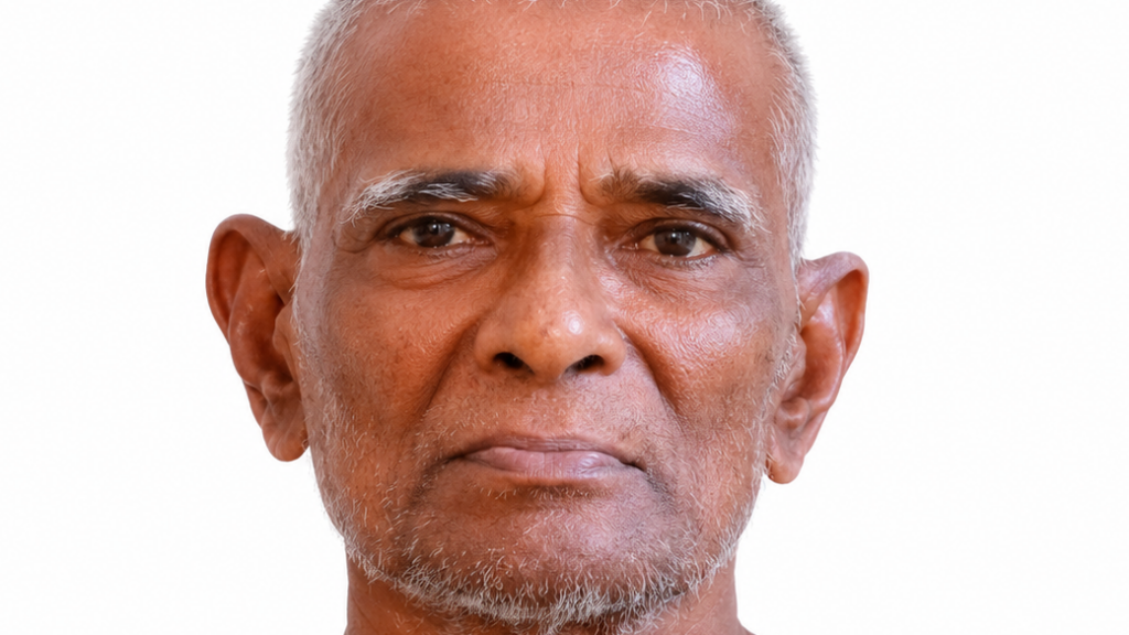 Close-up portrait of an older man with short gray hair and light stubble, facing the camera against a white background in a neutral expression.