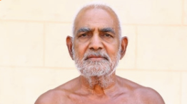 Portrait of an older man with a gray beard, bare shoulders, looking directly at the camera against a light, beige wall.