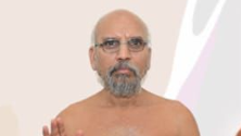 Older man with a gray beard and glasses, shirtless, facing the camera against a pale background with abstract shapes on the right.
