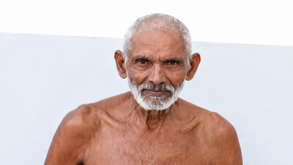 Older man with white beard and short white hair, shirtless, looking at the camera against a pale blue wall.