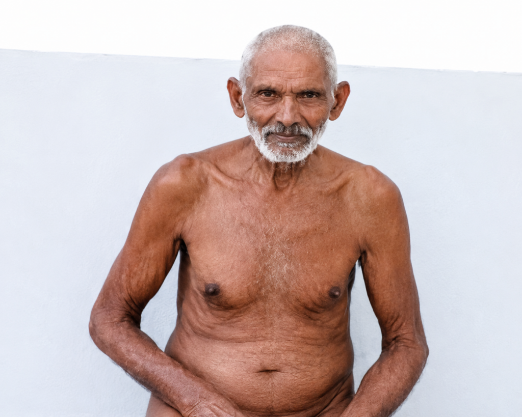 Older man with a gray beard, shirtless, facing the camera against a pale blue wall.