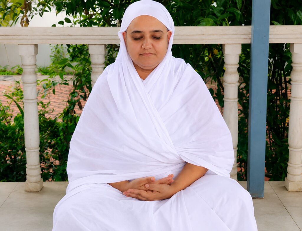 Person wearing white robes meditating with eyes closed, sitting cross-legged on a veranda with greenery in the background and railing behind.