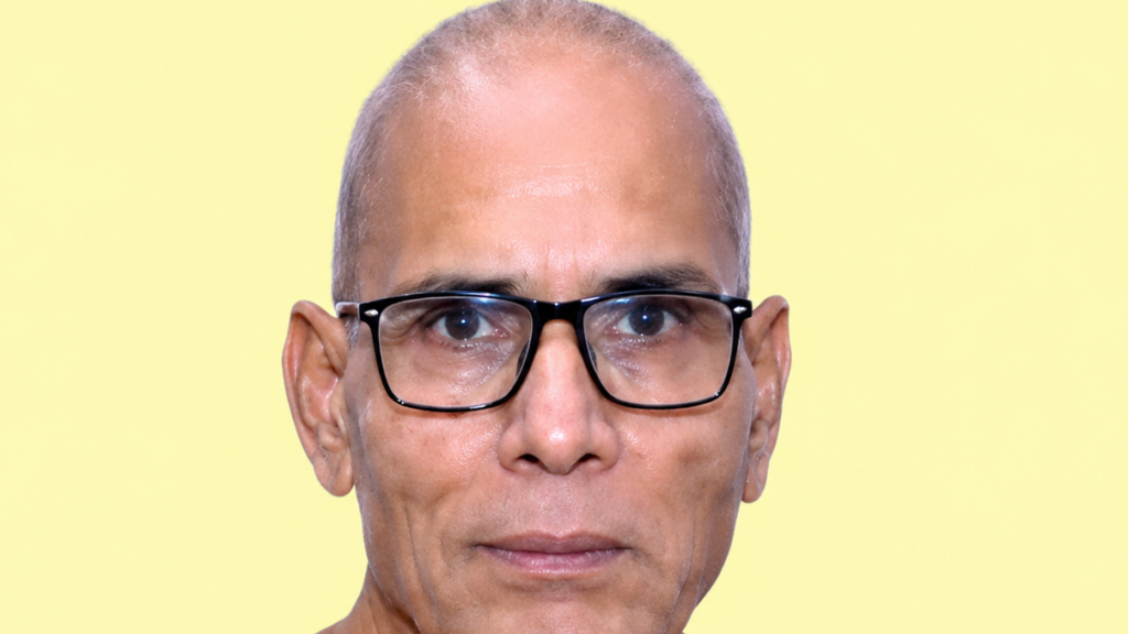 Close-up portrait of a middle-aged man with short gray hair and black rectangular glasses against a pale yellow background.