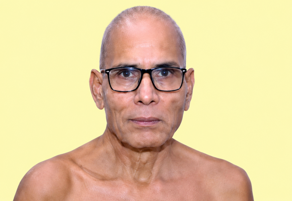 Close-up of a middle-aged man with black-framed glasses, bare shoulders, looking at the camera against a pale yellow background.