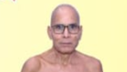 Older man with glasses, bald head and bare shoulders, neutral expression against a pale yellow background.