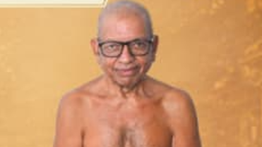 Older man with gray hair and black glasses, shirtless, standing against a gold background.