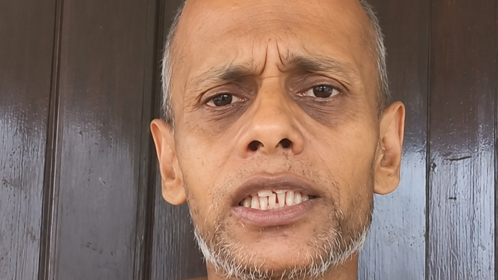 Close-up of a man with a furrowed brow and clenched teeth against a dark wooden background, conveying a tense expression.
