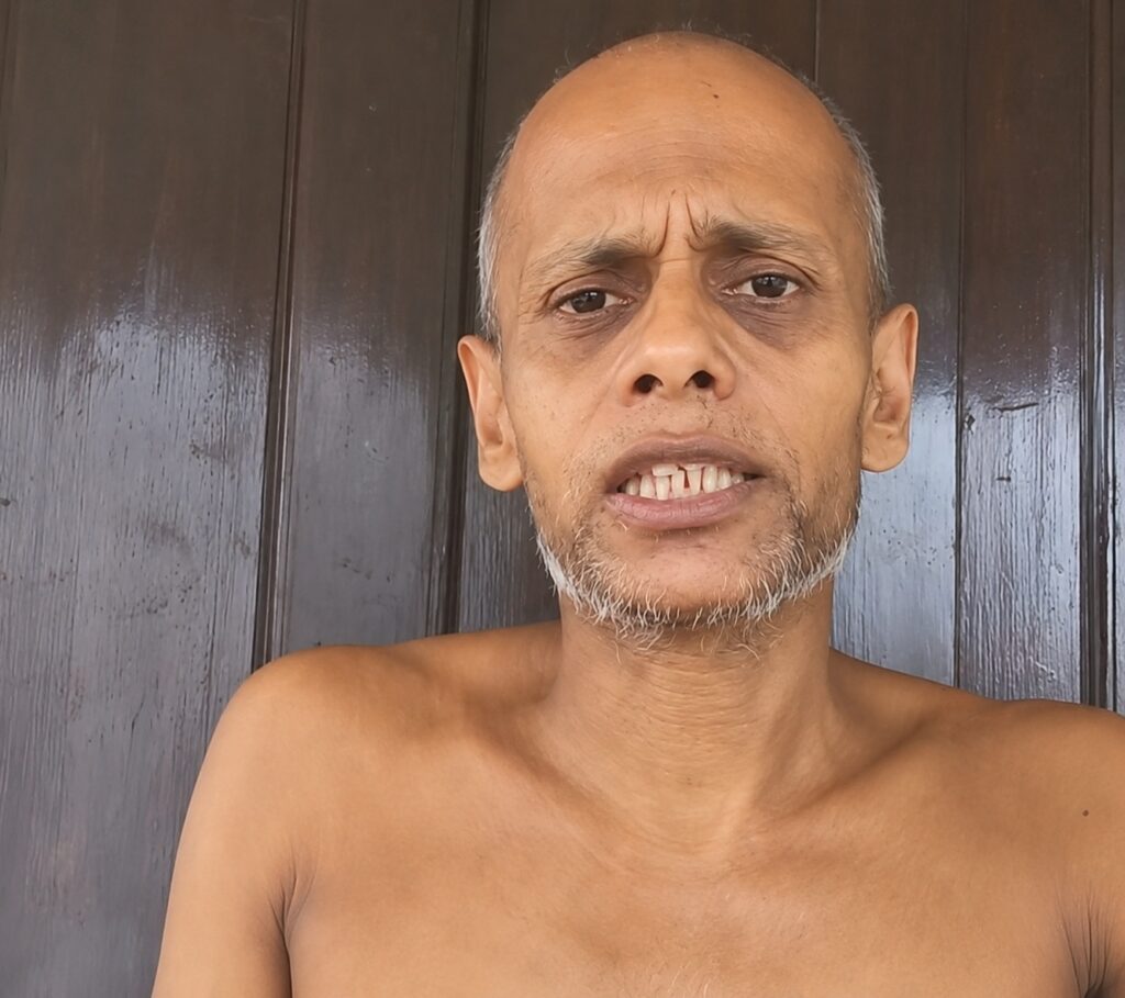 Shirtless middle-aged man with a gray beard looking at the camera, standing in front of a dark wooden background.