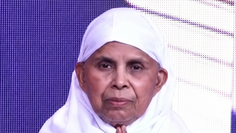 Elderly person wearing a white headscarf with hands pressed together in a prayer gesture.