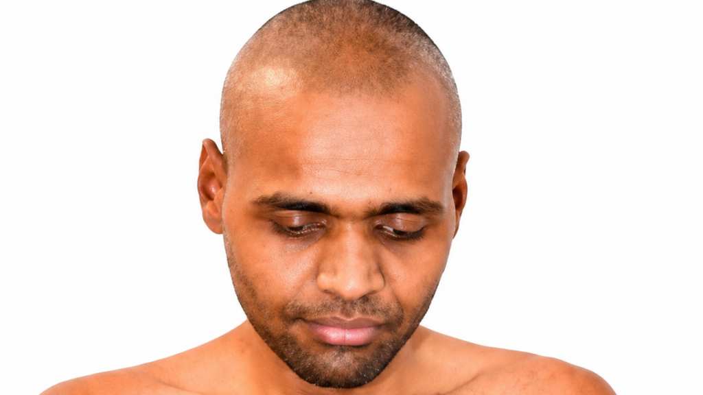 Close-up of a man with a shaved head looking downward, bare shoulders against a white background.