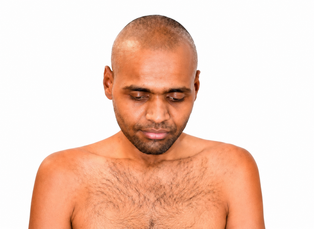 Man with a shaved head and chest hair looking downward against a white studio background.