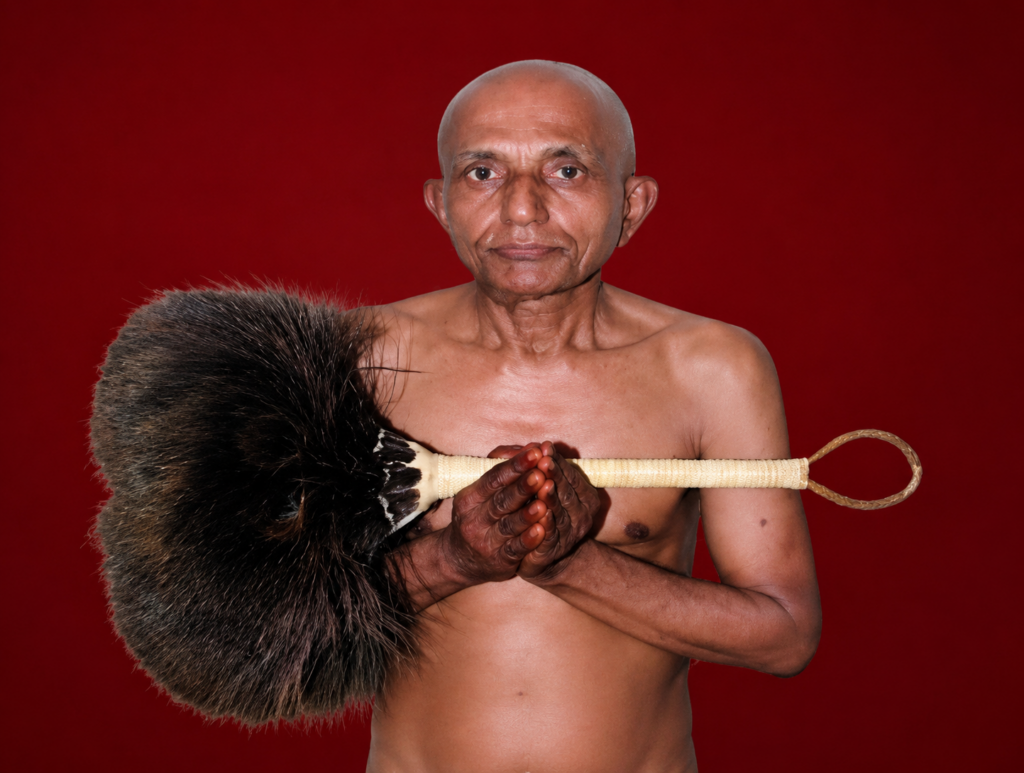 Senior bald man shirtless, holding a large feathered duster with a looped end across his chest, against a red background.