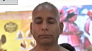 Man with a shaved head and eyes closed, calm expression, indoors with colorful, blurred background posters behind.