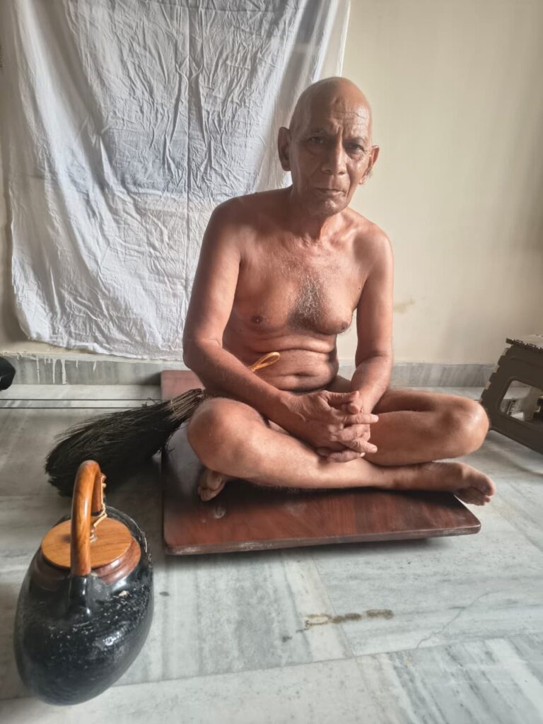 Elderly shirtless man sits cross‑legged on a wooden mat, hands folded in his lap in a simple room, with a kettle nearby.