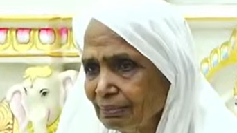 Close-up of an elderly woman wearing a white headscarf, looking solemn, with decorative elephant artwork behind her.