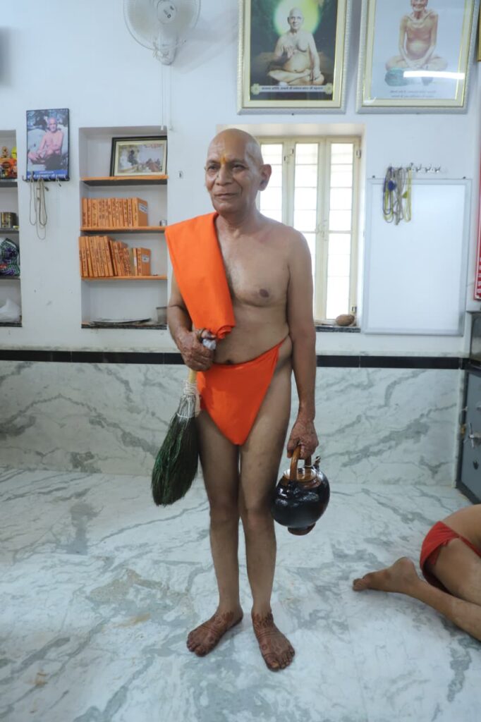 Elderly Hindu monk in orange robes stands barefoot in a temple room, holding a broom and a kalash for a ritual.