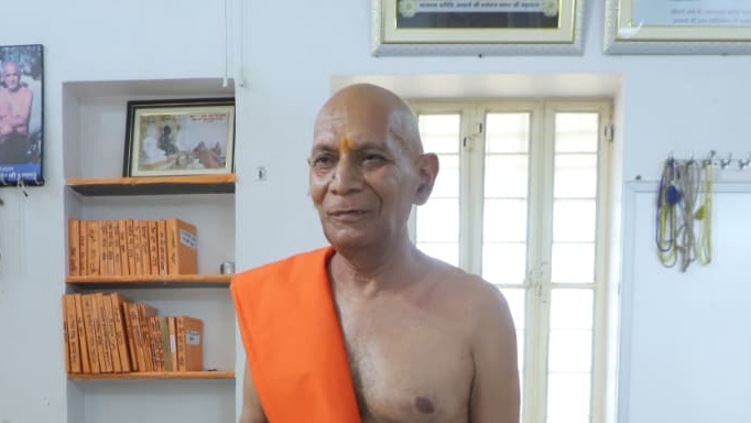Bald man in an orange robe stands indoors near a window, smiling, with orange boxes on shelves and framed photos on the wall.