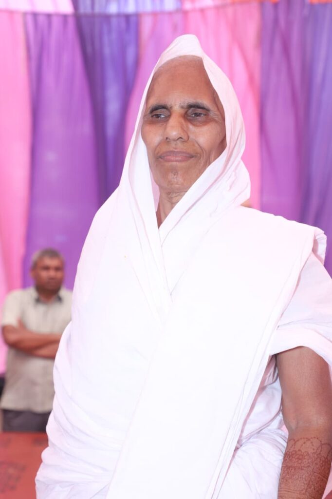 Person wearing a white traditional robe with a head covering at a colorful event backdrop of pink and purple drapes.