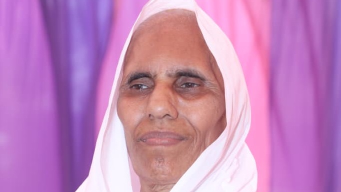 Older woman wearing a white hijab, smiling softly, with a purple-pink curtain background.
