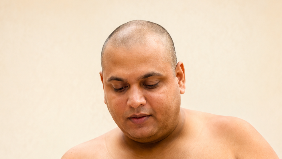 Shirtless man with a shaved head looking down against a beige background, close-up portrait.