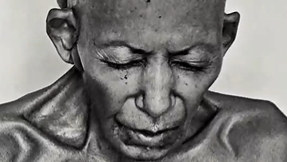 Close-up of a young boy with a shaved head and a somber expression, looking downward in grayscale.