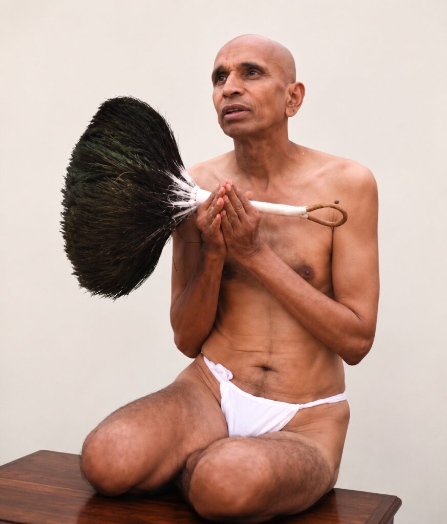 Shaven-headed man in a white loincloth sits on a wooden surface, holding a large black feather duster in a prayer pose.
