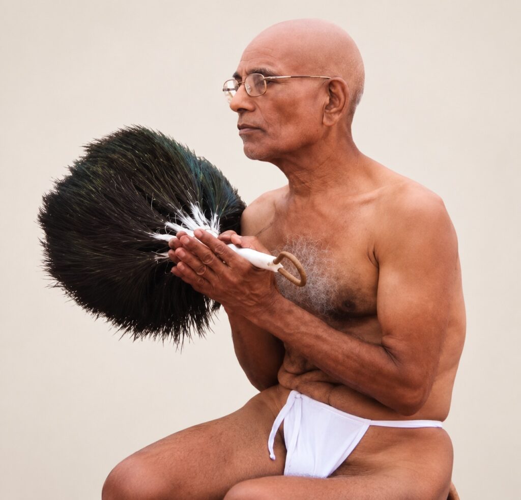 Older man with glasses, shirtless and in white briefs, holding a large black feather duster with both hands.