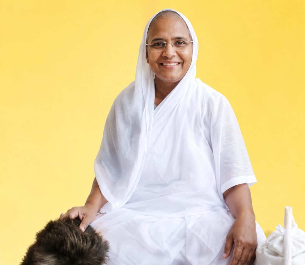 Smiling woman wearing a white hijab and robe, seated against a bright yellow background.
