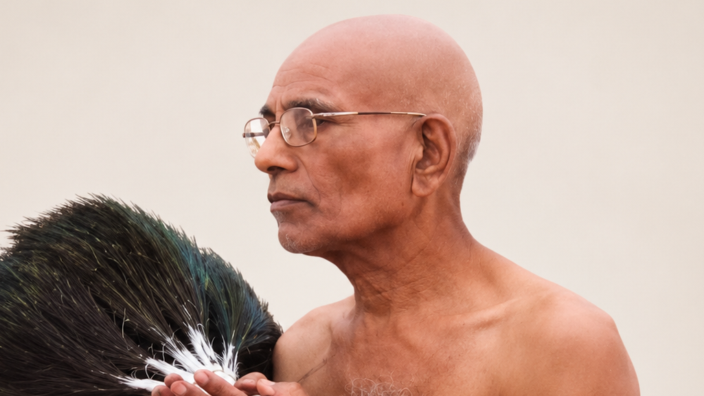 Bald man wearing glasses, shirtless, looking to the right with a large peacock feather beside him against a light background.
