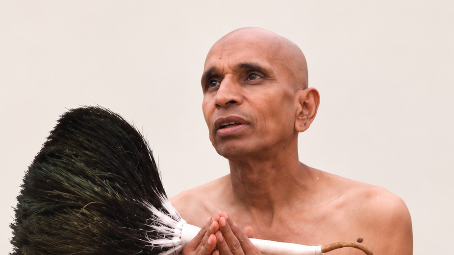 Bald man with bare shoulders clasps his hands in prayer while holding a large broom with dark bristles against a plain background.
