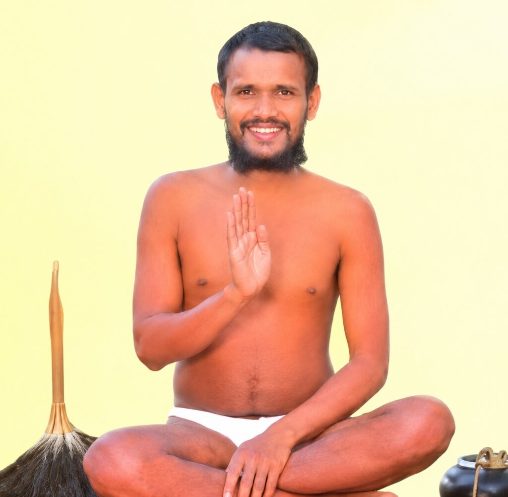 Smiling, shirtless man sits cross-legged on a pale yellow background, raising his right hand in a blessing gesture, with a broom and a pot nearby.