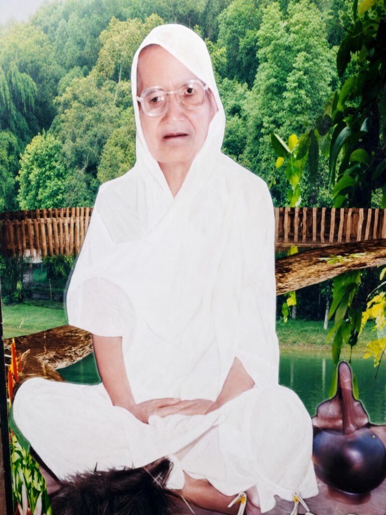Senior woman in a white robe and headscarf, wearing glasses, seated cross‑legged outdoors among greenery.