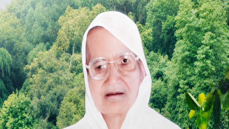 Portrait of an elderly woman with a white headscarf and glasses, standing in front of a green forest background