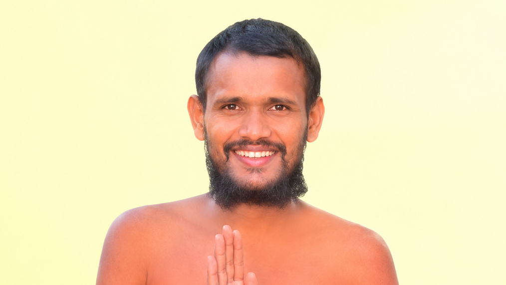 Smiling man with a short beard, hands pressed together in a Namaste greeting, against a pale yellow background.