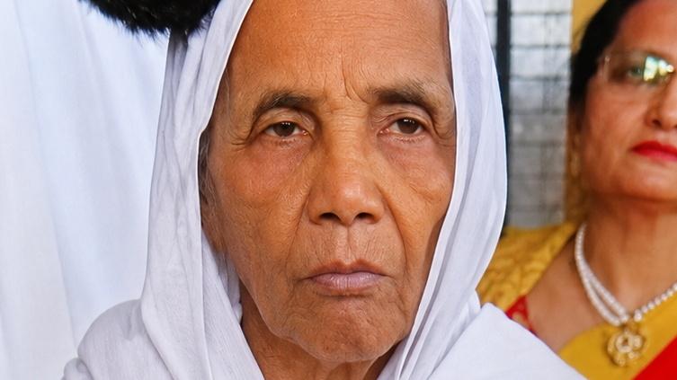 Close-up of an elderly woman in a white headscarf with a serious expression, background shows a woman in yellow attire and glasses.
