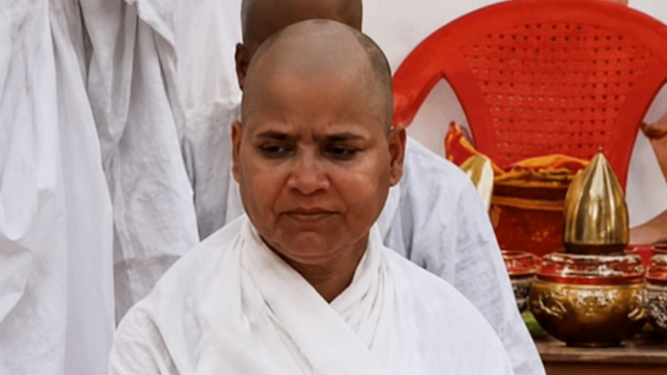 Buddhist monk with a shaved head in white robes, sitting indoors with a red chair and ceremonial brass bowls nearby.
