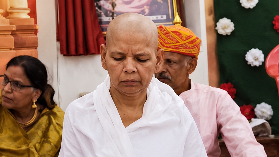 Buddhist monk with a shaved head wearing white robes, eyes down in ceremony, with attendees in traditional attire in the background.