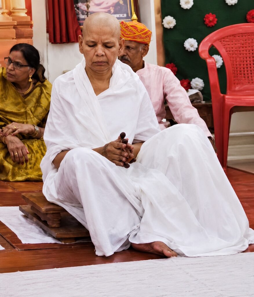 Monk in white robes meditating with clasped hands, seated on a wooden mat in a temple setting