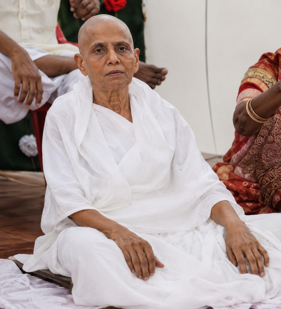 Elderly person with a shaved head in white robes seated cross‑legged at a ceremony, calm expression.