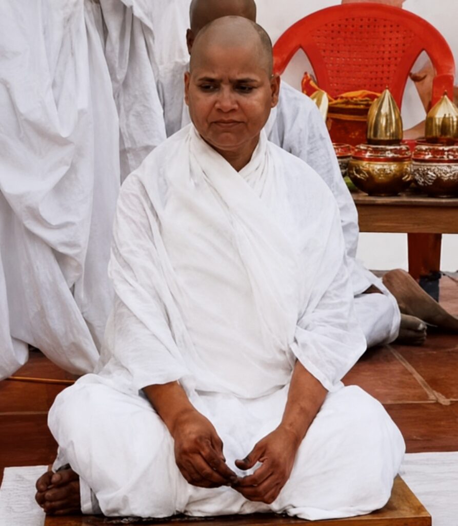 Meditating Buddhist monk in white robes seated cross‑legged on a wooden floor, hands resting in lap, with ceremonial items nearby