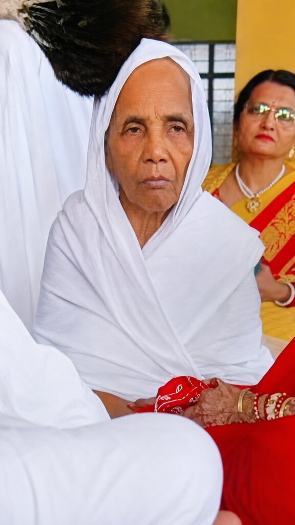 Older Indian woman in a white sari looking somber, seated with hands resting in her lap during a ceremony, nearby people in colorful traditional attire in the background