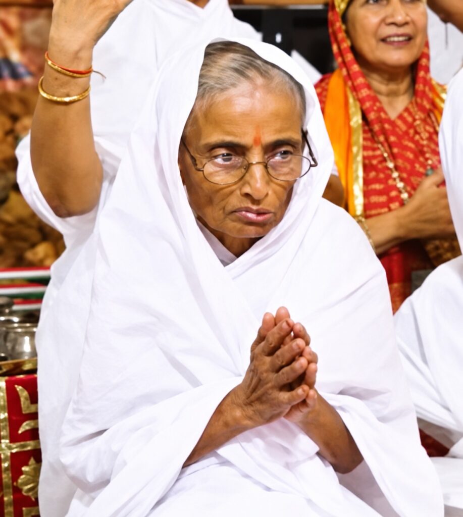 Elderly Indian woman in a white saree with glasses, hands pressed together in prayer, red tilak on her forehead.