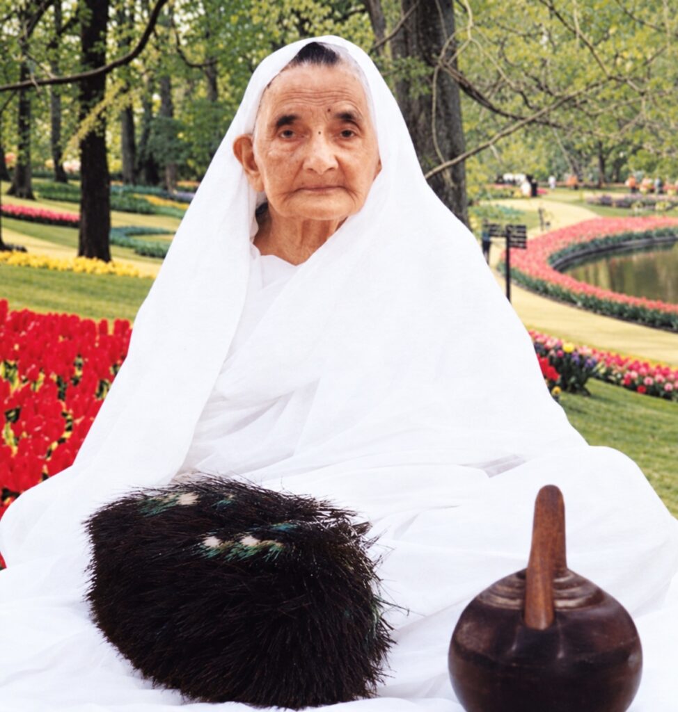 Portrait of an elderly woman in a white headscarf sitting in a colorful garden with flowers and trees in the background.