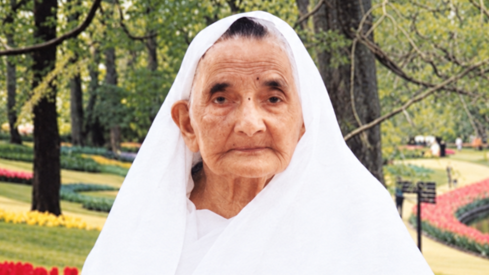 Elderly woman in a white veil standing in a park with colorful flower beds in the background, looking at the camera.