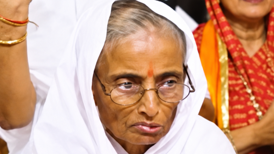 Close-up of an elderly woman wearing a white headscarf and glasses at a cultural or religious gathering, looking thoughtful.